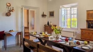Zona pranzo con tavolo in legno apparecchiato per un pasto, una finestra vista verde e una cucina rustica sullo sfondo.