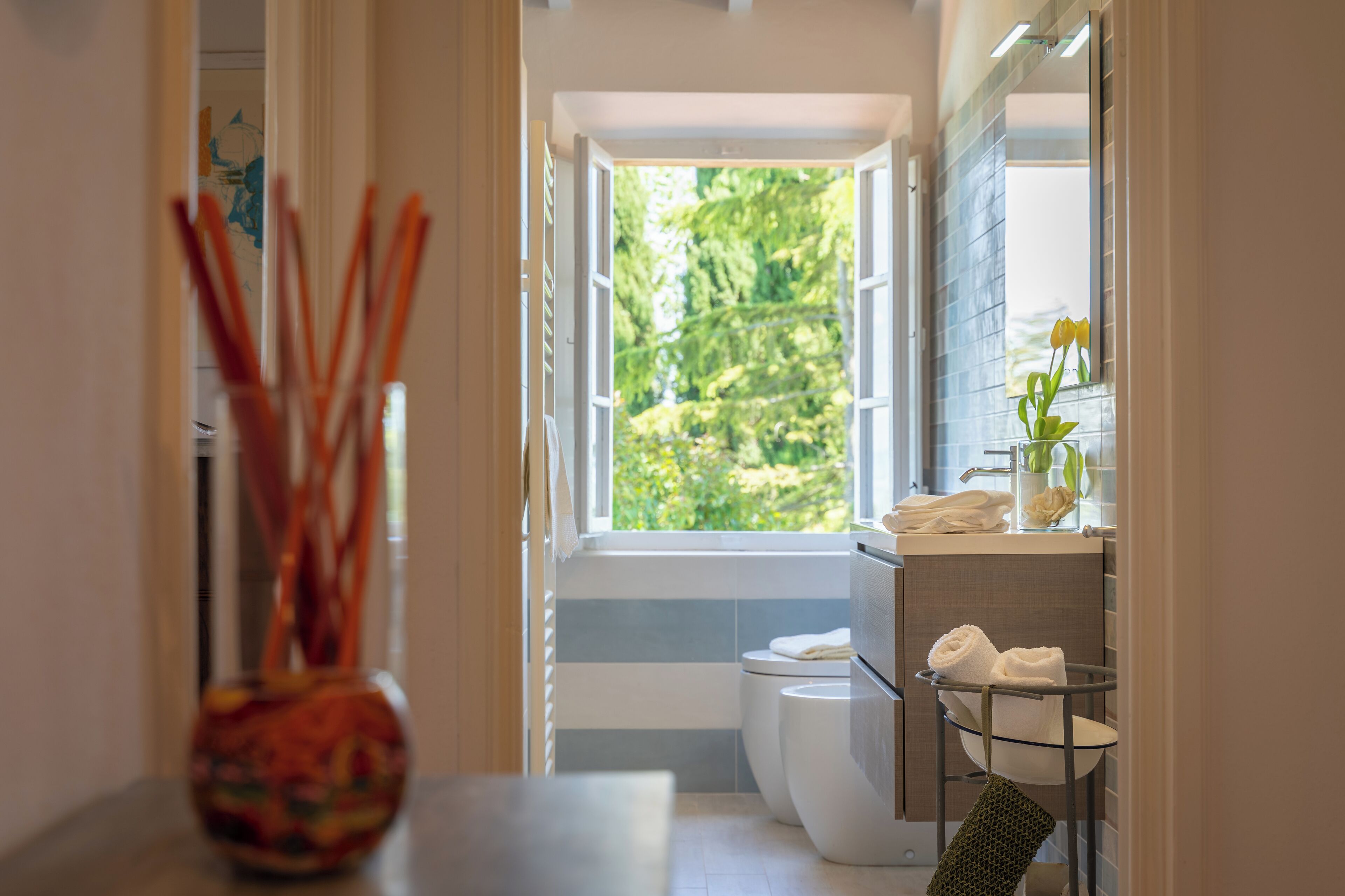 Un baño con ventana, inodoro, lavamanos y un florero con ramitas de caña.