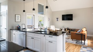 A modern kitchen with white cabinets, a black countertop, and pendant lights. There is a dining area with a brown leather couch and a flat-screen TV mounted on the wall.