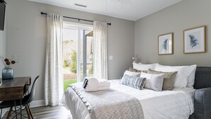 A bedroom with a bed, bedside table, and chair. There is a window with curtains, a door, and two framed pictures on the wall.