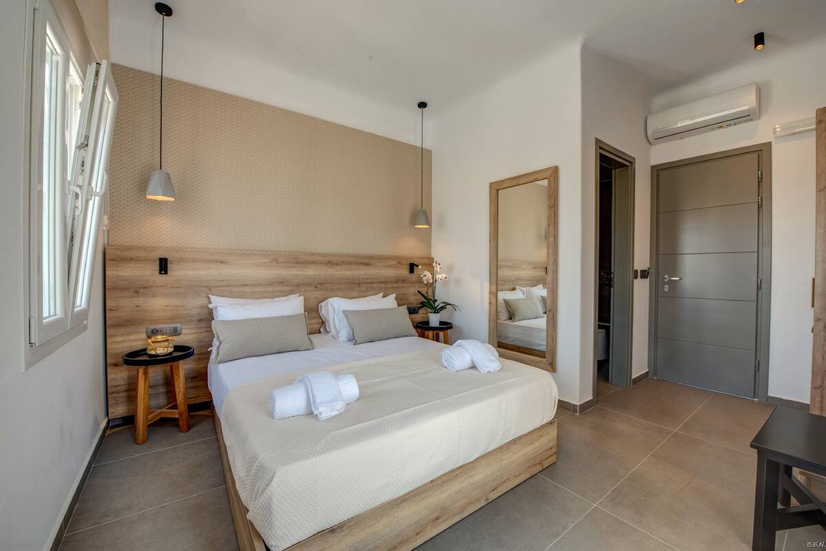 A modern hotel room with a large bed, wooden headboard, bedside tables, a mirror, and an air conditioning unit.