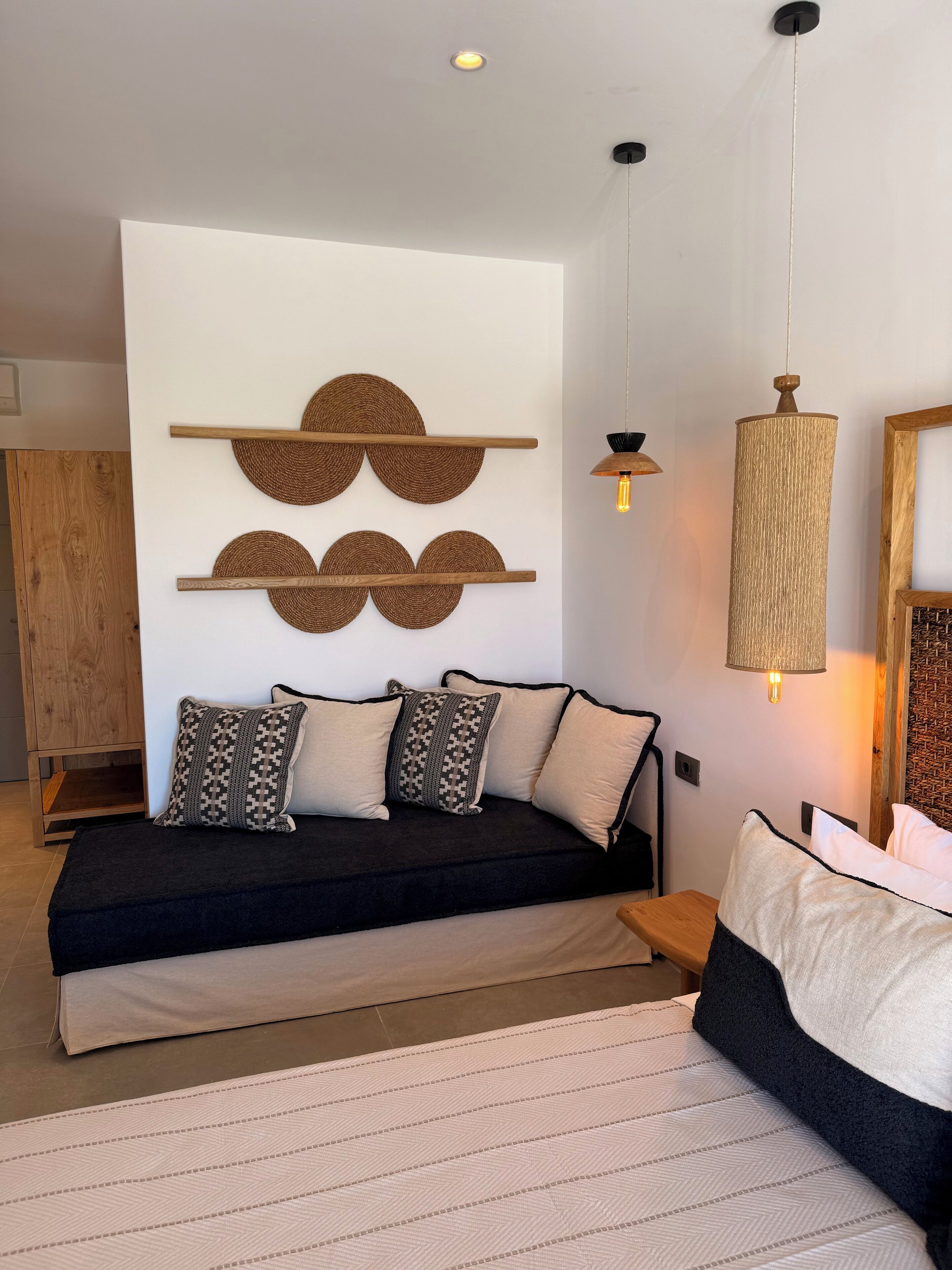 Ein modernes Schlafzimmer mit einer dunkelgrauen Couch, zwei gemusterten Kissen, einer gewebten Wanddekoration, einer hängenden Leuchte und einem Holznachttisch.
