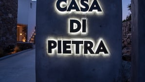 Ein Schild mit der Aufschrift 'CASA DI PIETRA' mit beleuchteten Buchstaben, vor einer Fassade aus Steinwänden und einem Hauseingang.