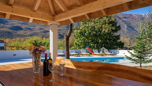 A wooden table with a bottle of wine and glasses, overlooking a pool with lounge chairs and a mountain view. - Lovely home in Zagvozd (Zagvozd)