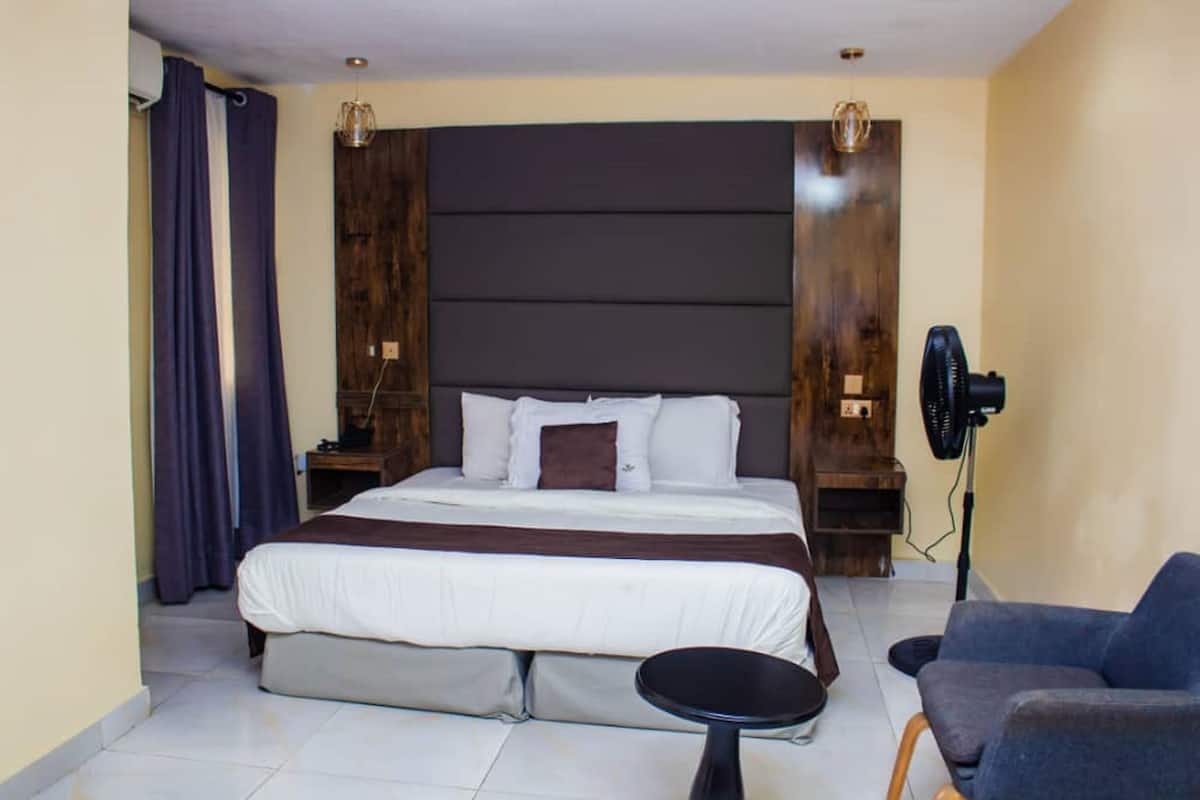A bedroom with a large bed, a bedside table, a chair, a standing fan, and a wall-mounted light fixture.