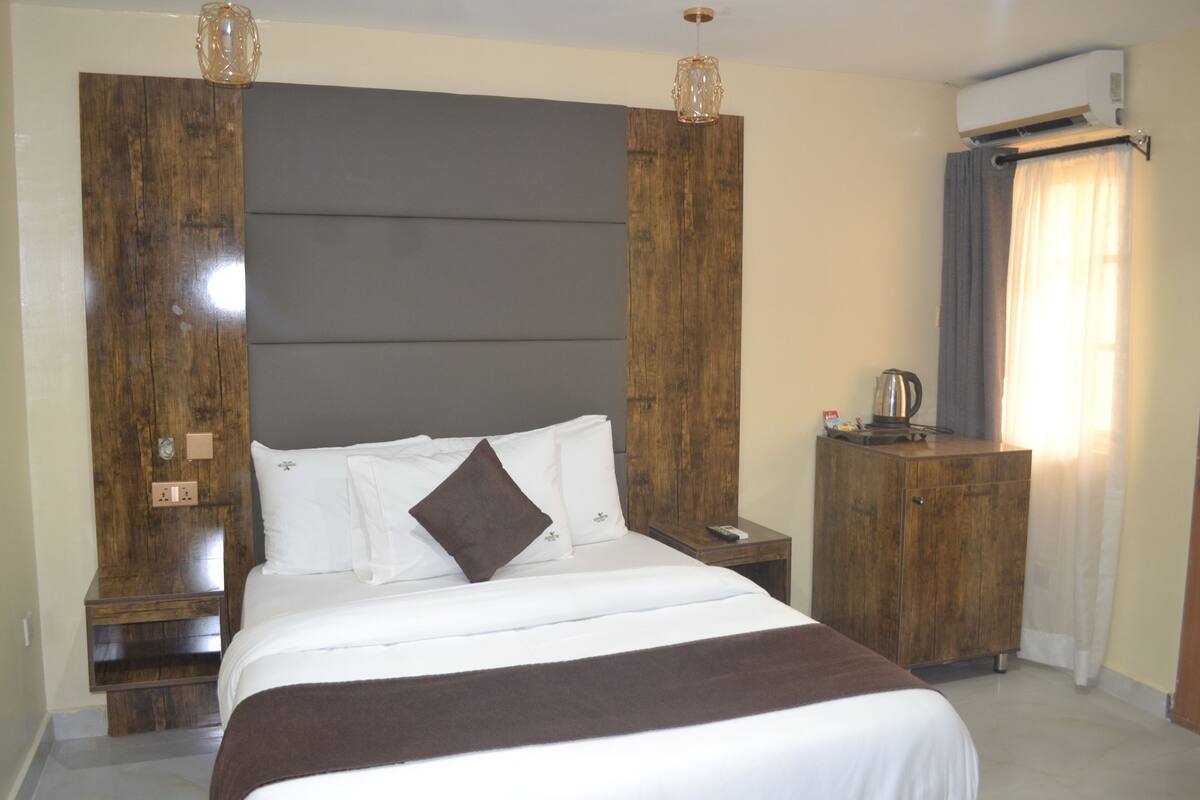 A hotel room with a large bed, wooden headboard, bedside tables, a wardrobe, and a window with curtains.