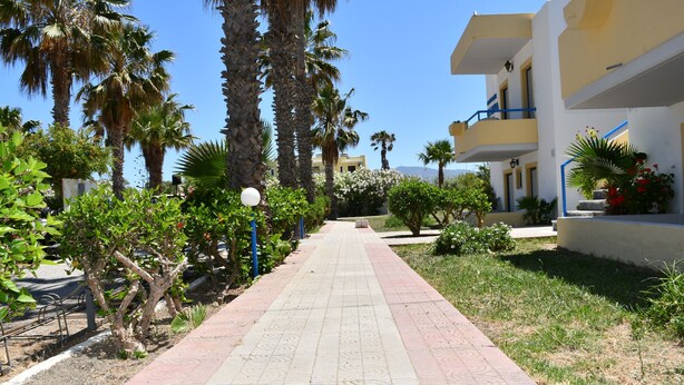 A pathway lined with palm trees and greenery, leading to a building with balconies and a clear blue sky. - 1-bedroom Kos Olympia Apartment at Lambi Beach (Kos)