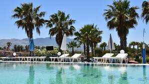 A pool area with lounge chairs and umbrellas, surrounded by palm trees and mountains in the background. - 1-bedroom Kos Olympia Apartment at Lambi Beach (Kos)