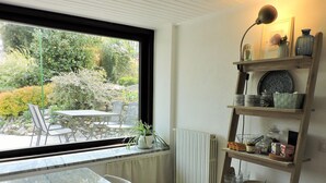A room with a dining table and chairs, a window with a view of greenery, a radiator, and a wooden shelf with various items. - Spending a vacation in the Garden. (Barzola)