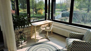A sunlit room with a desk, chair, and a view of a garden through large windows. - Spending a vacation in the Garden. (Barzola)