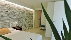 A bedroom with a white bed, a stone wall, a bedside table with a lamp, and a door leading to another room. - Spending a vacation in the Garden. (Barzola)