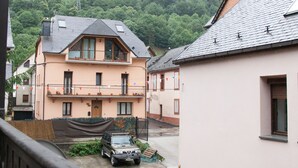 Un immeuble résidentiel doté d’un balcon, avec une voiture garée devant et une vue sur une colline boisée.