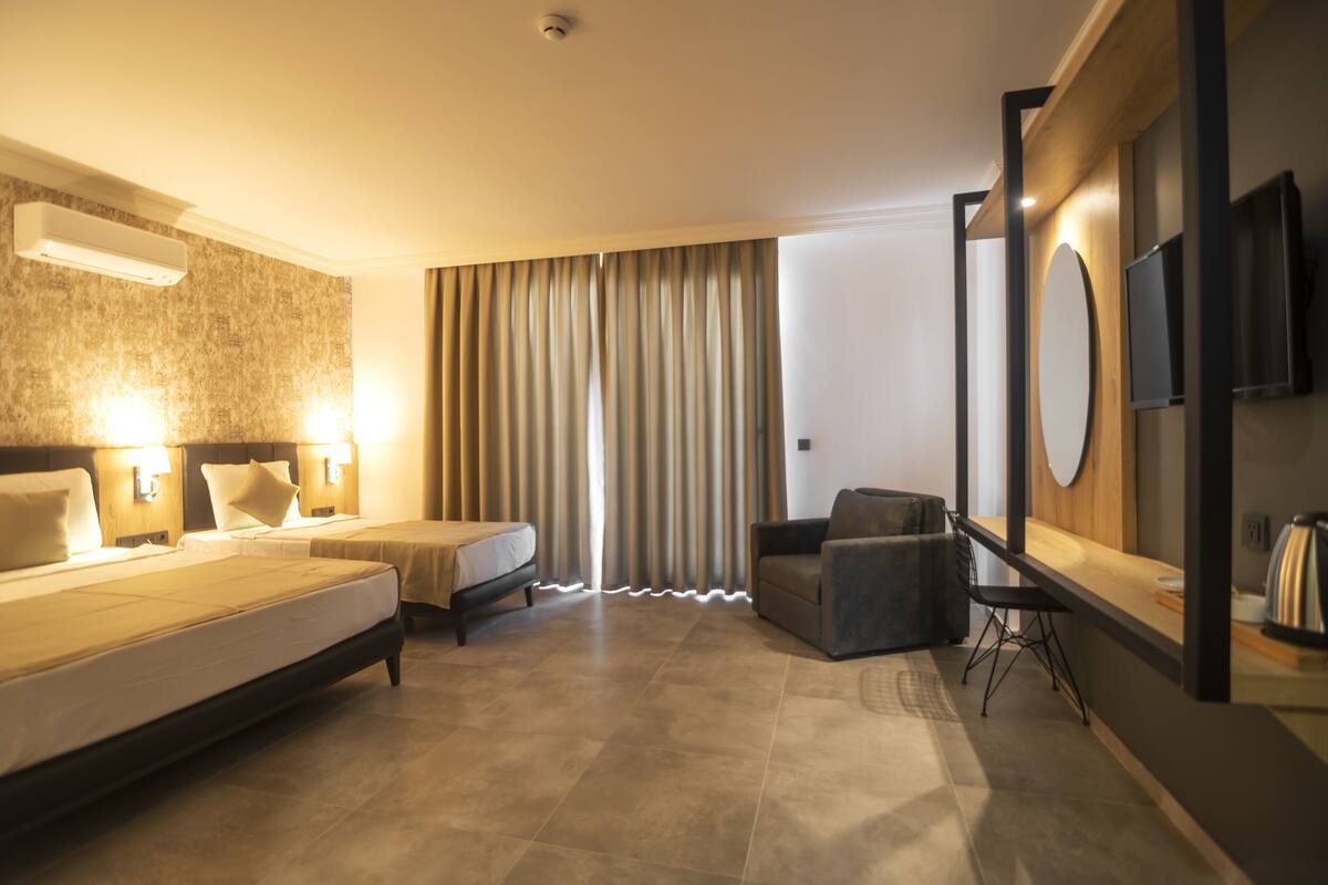 A modern hotel room with a large bed, two bedside tables with lamps, a sofa, a small table, and a TV mounted on the wall.