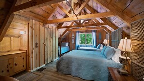 A rustic bedroom with a wooden ceiling, a bed with a blue bedspread, a nightstand, and a window with curtains.