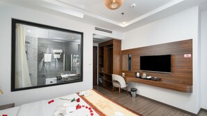 Una habitación de hotel moderna con una cama grande, un televisor montado en la pared, un escritorio con una silla y una zona de ducha visible a través de una puerta de cristal.