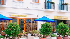 Un hotel con cafetería y restaurante en la planta baja, mesas exteriores con sombrillas y plantas en macetas.