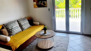 A living room with a yellow sofa, patterned cushions, a small wooden table with a plant, and a sliding glass door leading to an outdoor area with a white fence.