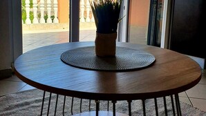 A round wooden table with a woven placemat and a potted plant in the center, positioned in a room with large glass doors leading to an outdoor area with a view of a hilly landscape.