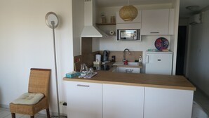 A compact kitchen with white cabinets, a microwave, a sink, and a small refrigerator. There is a wicker chair with a cushion and a floor lamp. - Sol-y-Days le Mayflower, Apartment for 6 people with swimming pool (Le Grau-du-Roi)