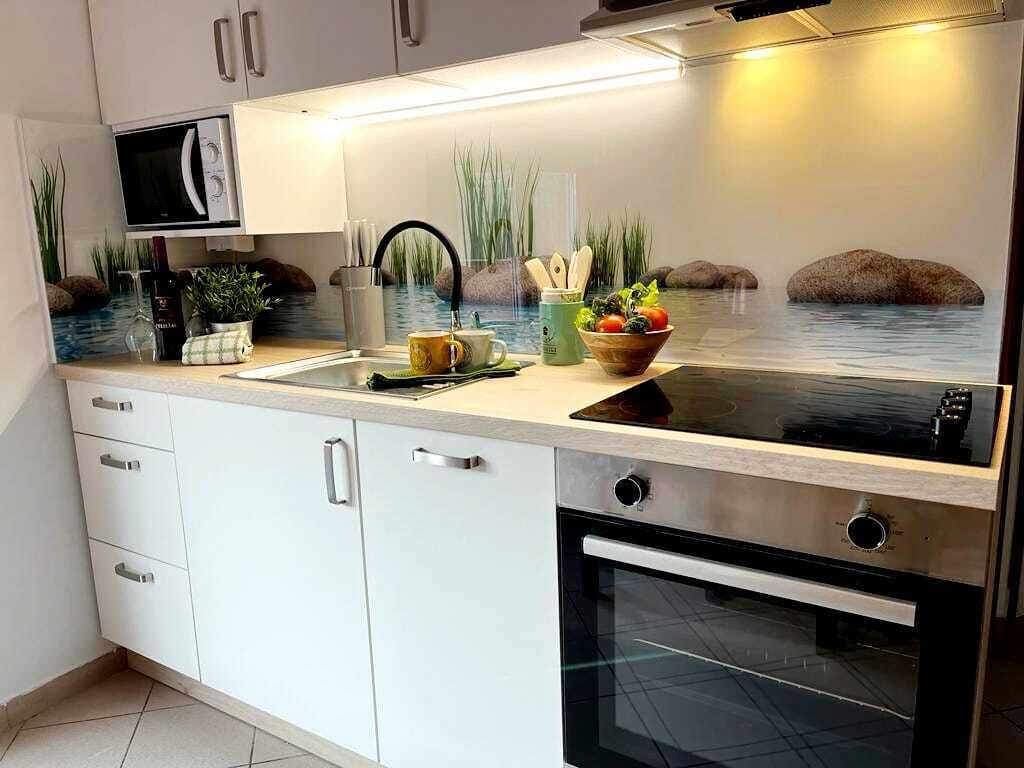 Une cuisine moderne avec des armoires blanches, un four à micro-ondes encastré, une plaque de cuisson à induction noire et un four en acier inoxydable. On y trouve un évier avec un robinet, un panier de fruits et une bouteille de vin sur le comptoir.