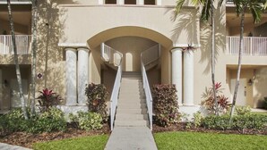 A building entrance with a central staircase flanked by columns, surrounded by well-maintained landscaping and palm trees. - Beautiful Condo with a Tree House Feel (Port St. Lucie)