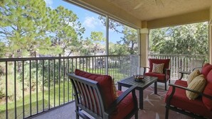 A covered outdoor patio with a table and two red chairs, overlooking a well-maintained garden and trees. - Beautiful Condo with a Tree House Feel (Port St. Lucie)