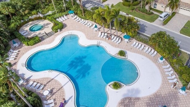 A large, kidney-shaped swimming pool surrounded by lounge chairs and umbrellas, with palm trees and a residential area in the background. - Beautiful Condo with a Tree House Feel (Port St. Lucie)