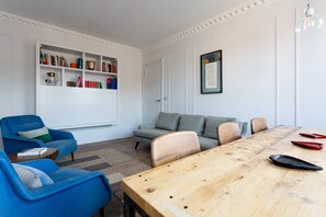 Una sala de estar moderna con una mesa de comedor de madera, sillones azules, un sofá gris y una estantería llena de libros y objetos decorativos.