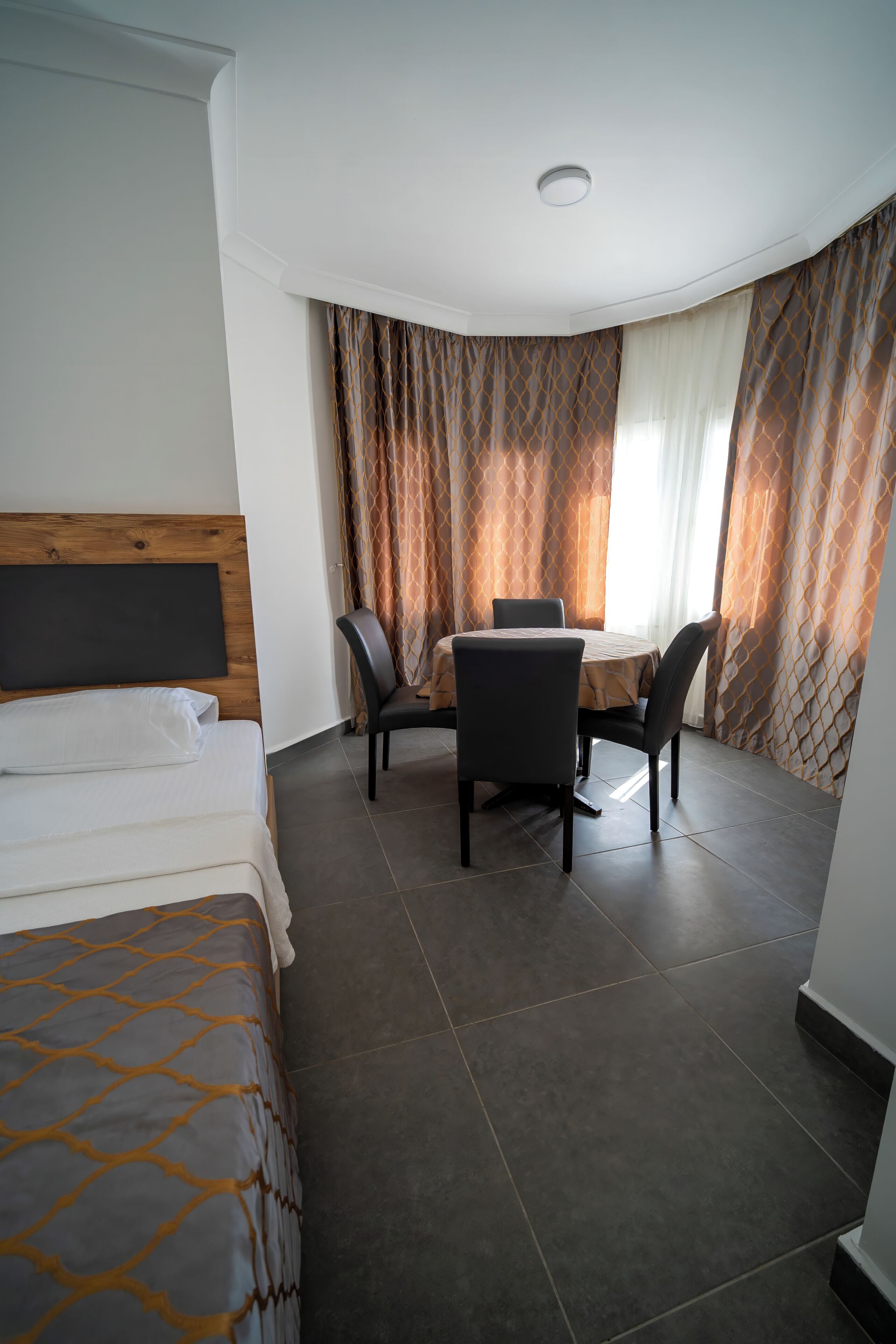 A hotel room with a bed, a dining table with chairs, a window with curtains, and a ceiling light.