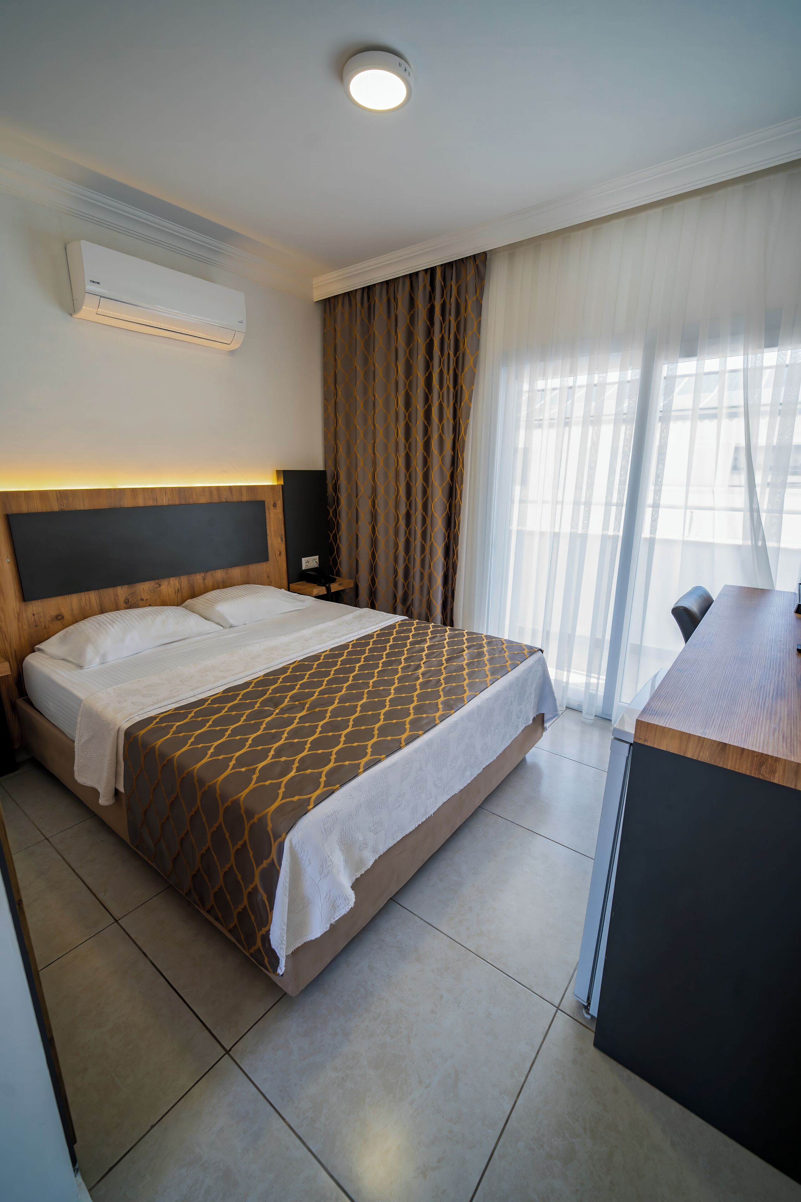 A hotel room with a bed, a desk, an air conditioner, and a view through sheer curtains.