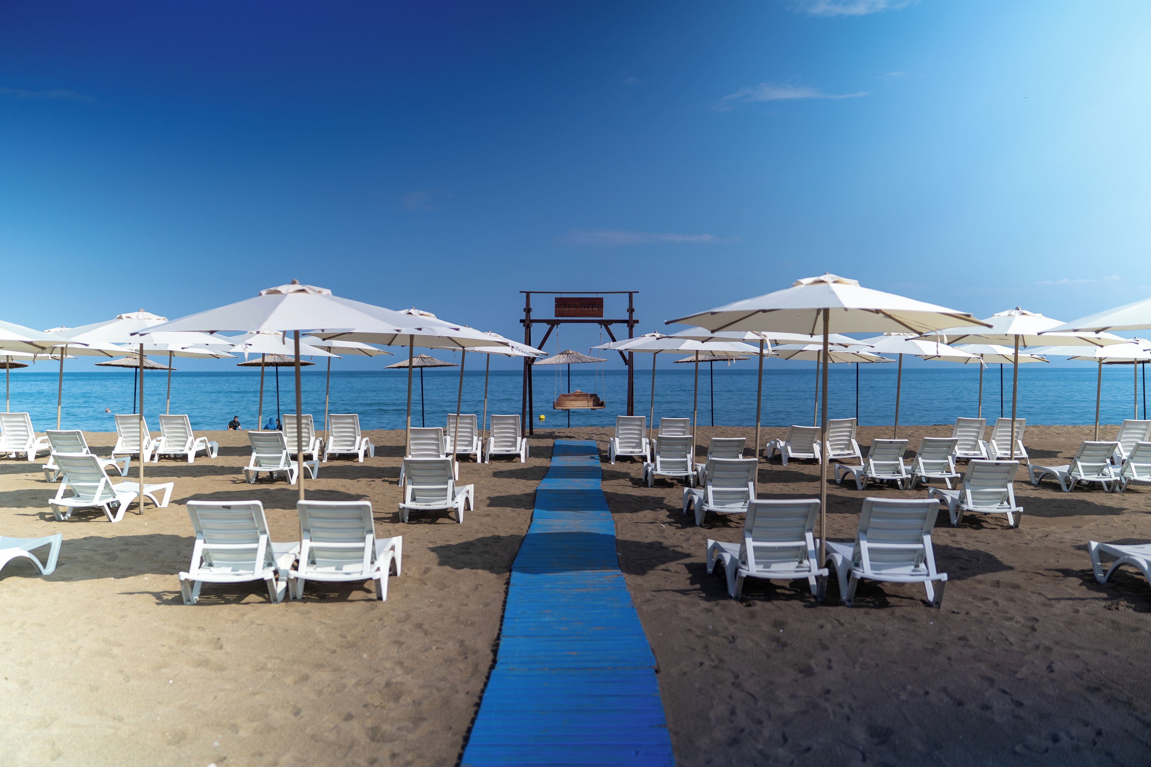 A beach with rows of white lounge chairs and umbrellas, a blue pathway leading to a wooden structure, and the ocean in the background.