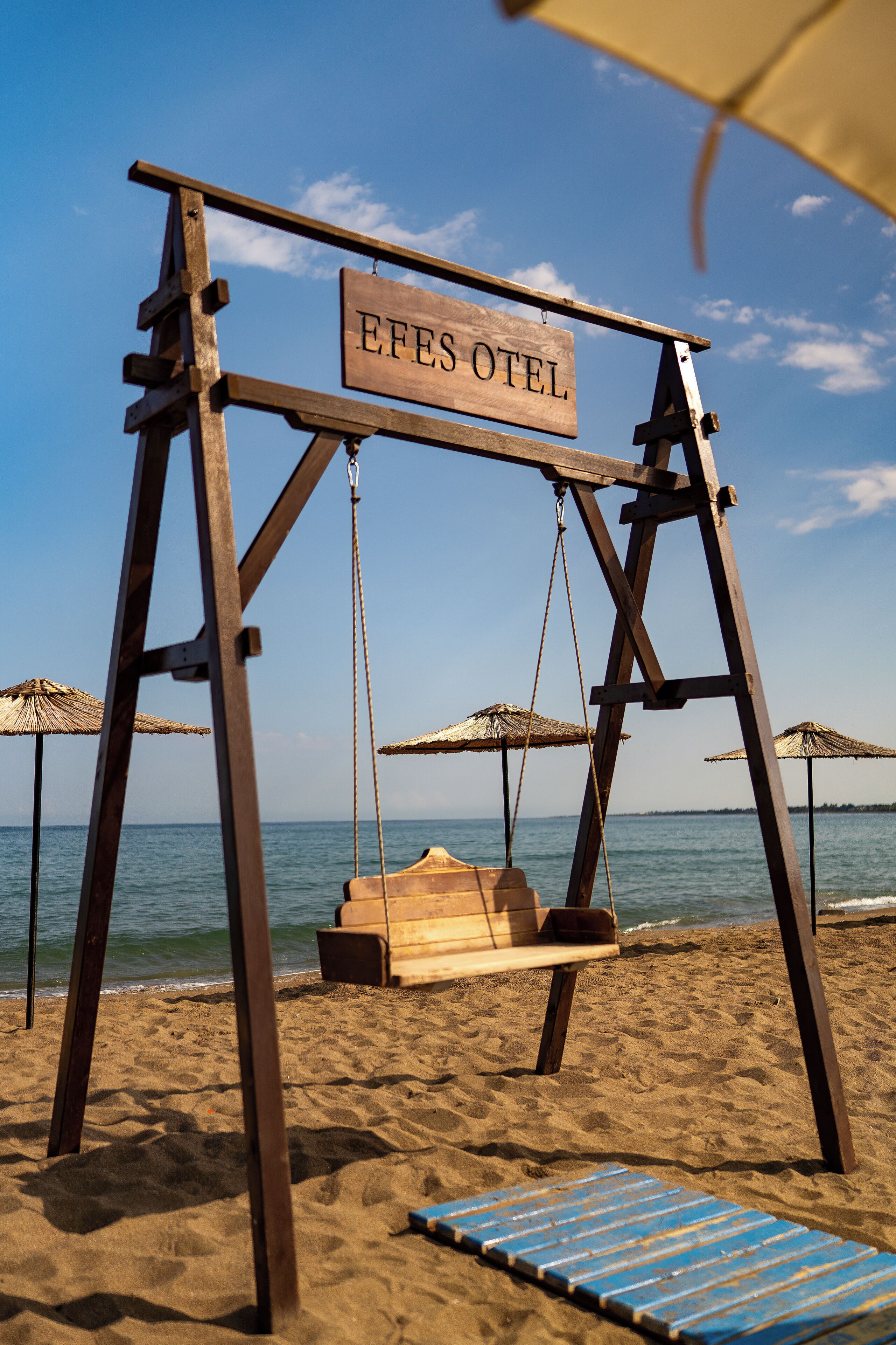 A wooden swing set with a sign that reads 'EFES OTEI' on a sandy beach with umbrellas and a view of the sea.