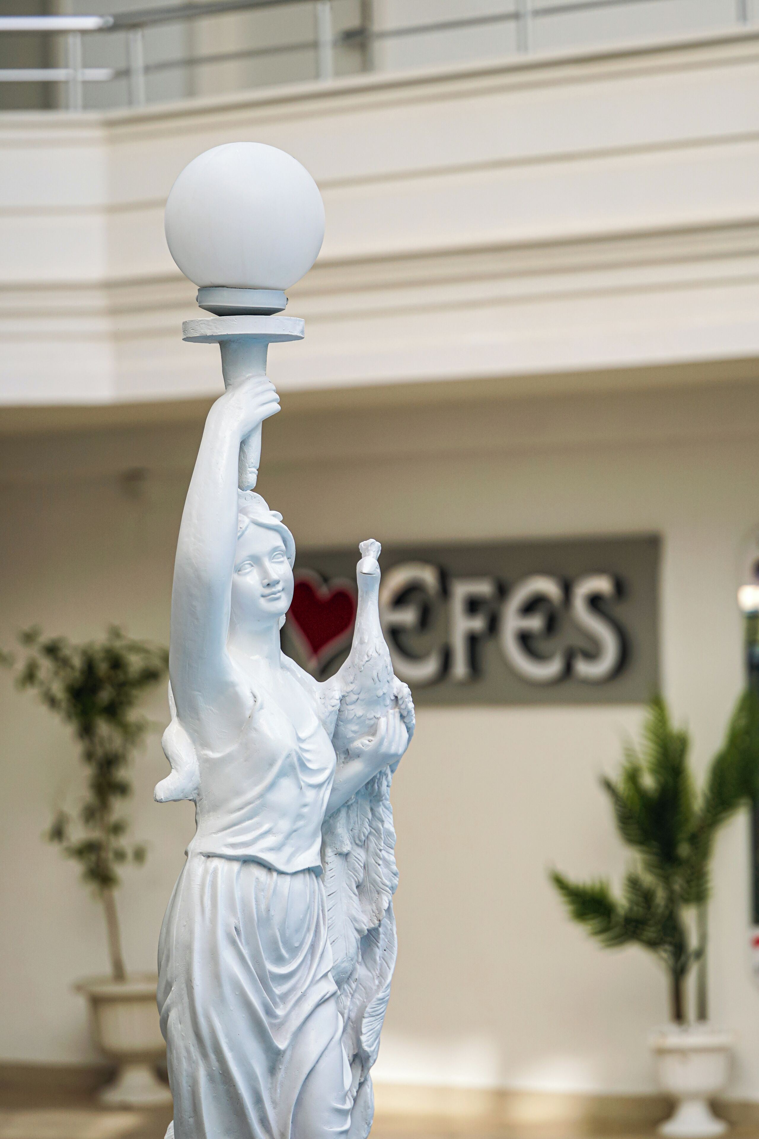 Una estatua blanca en un espacio público interior, con un cartel de fondo.