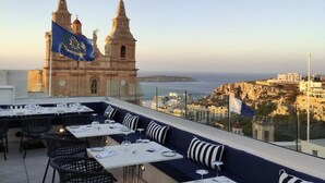 Un espacio para comer al aire libre con mesas y sillas, con vistas a una ciudad costera y una iglesia histórica con dos torres.