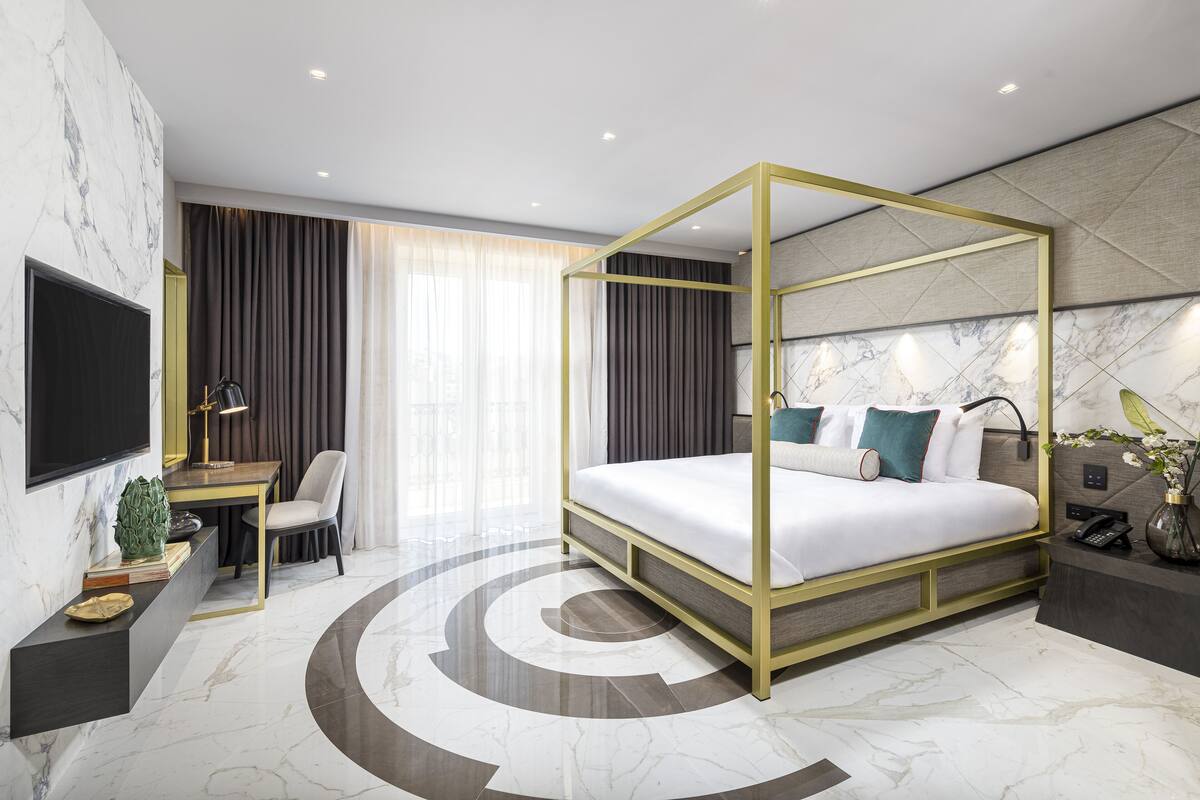 A modern hotel room with a four-poster bed, a desk with a chair, a flat-screen TV, and a large window with curtains.