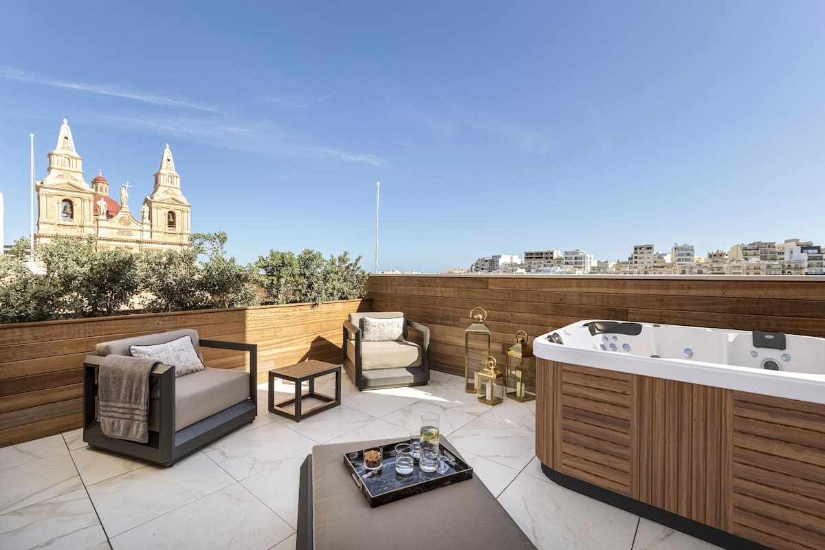 A rooftop terrace with a sofa, chairs, a hot tub, and a tray with glasses and a bottle.