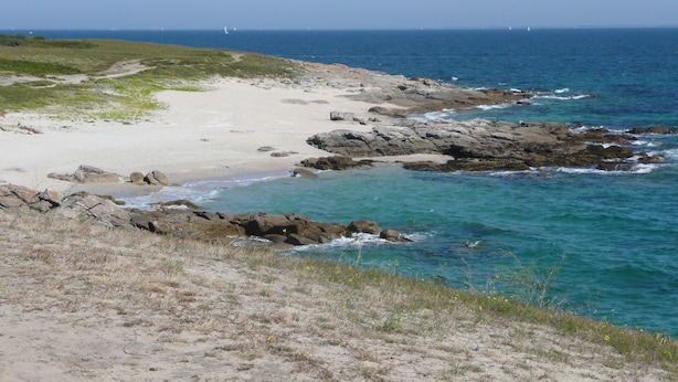 Ett kustlandskap med en sandstrand, en klippig kust och ett klart, blått hav.