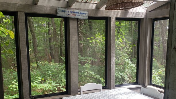 A rustic wooden room with a table and chairs, large windows offering a view of a forest, and a hanging light fixture.