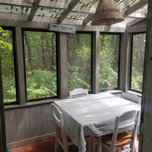 Een rustieke houten kamer met een tafel en stoelen, grote ramen met uitzicht op een bos, en een hanglamp.