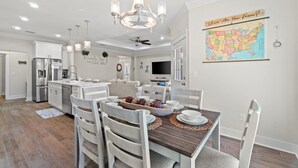 A modern kitchen with white cabinetry, a dining area with a wooden table, and a map on the wall. - Serenity Tides PCB- Golf Cart, Games, MiniGolf & Dogs OK – 3 Blocks to Beach (Panama City Beach)