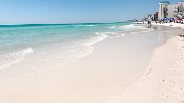 A beach with clear turquoise water and a sandy shore. There are buildings in the background, and people are visible on the beach. - Serenity Tides PCB- Golf Cart, Games, MiniGolf & Dogs OK – 3 Blocks to Beach (Panama City Beach)