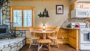 A cozy cabin kitchen with a stone fireplace, wooden dining table, and a bear-themed wall decoration. - Walk to Lake Cabin great for kids &  Pets Spa BBQ WiFi King Bed Ski Free Coffee (Big Bear Lake)