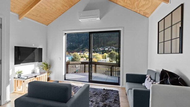 Un salon moderne doté d’une grande porte-fenêtre coulissante donnant sur un balcon, d’un téléviseur à écran plat fixé au mur et d’un coin salon confortable avec deux canapés.