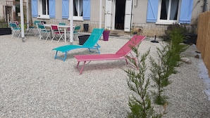 A covered outdoor seating area with lounge chairs and a table, a stone building with blue shutters, and a gravel ground.