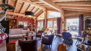 A cozy log cabin interior with a wooden ceiling, a sofa, multiple chairs, a table with drinks, and a view of the outdoors.