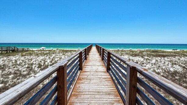 Une promenade en bois qui mène à une plage où l’eau est d’un bleu limpide et le rivage est sablonneux.