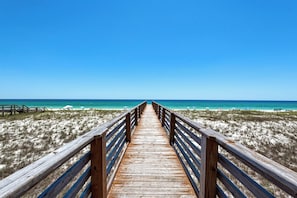 A wooden boardwalk leading to a beach with clear blue water and a sandy shore.