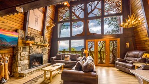 A cozy lodge interior with a stone fireplace, leather furniture, and large windows offering views of the outdoors. - Stunning view, location & Cabin amenities- 2.3 mi to Gatlinburg (Gatlinburg)