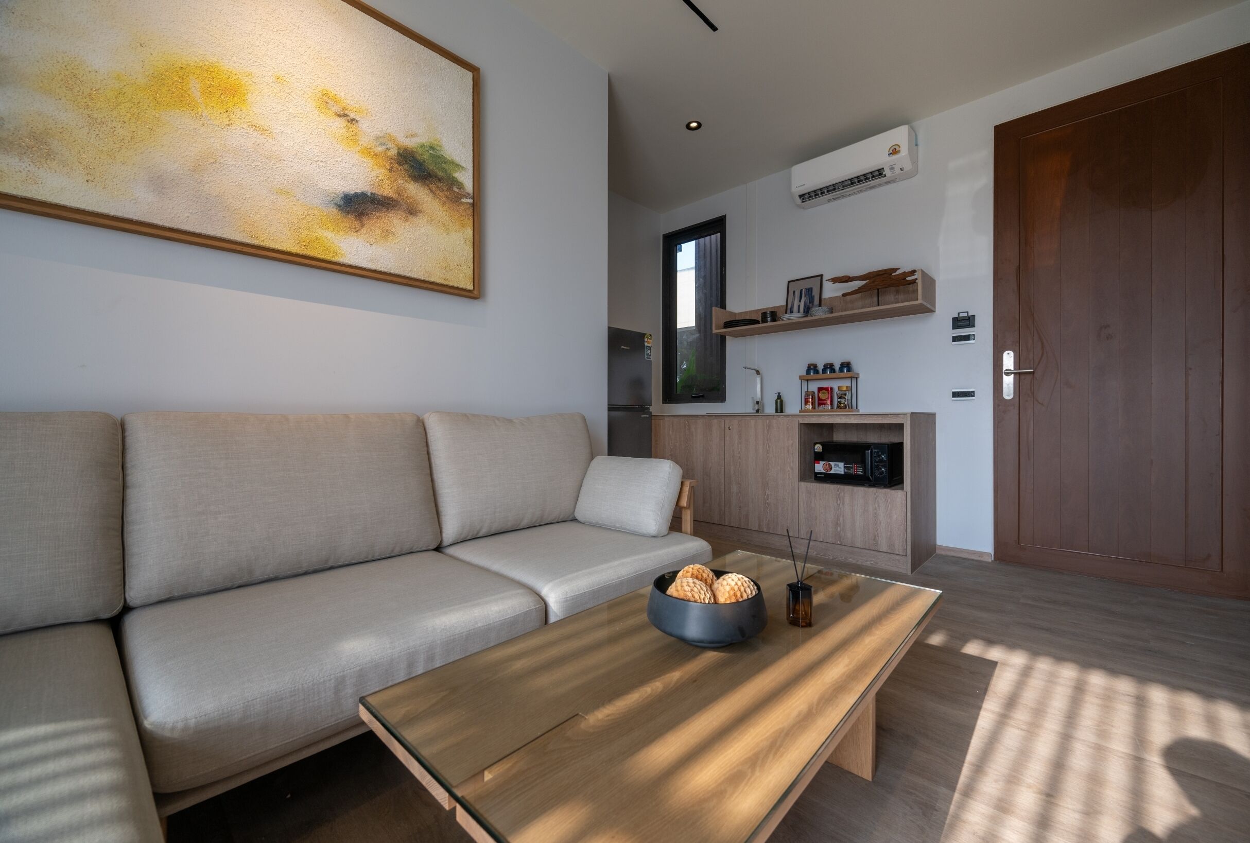 A modern living room with a sofa, a coffee table with decorative items, a kitchen area with a refrigerator, and a wooden door.