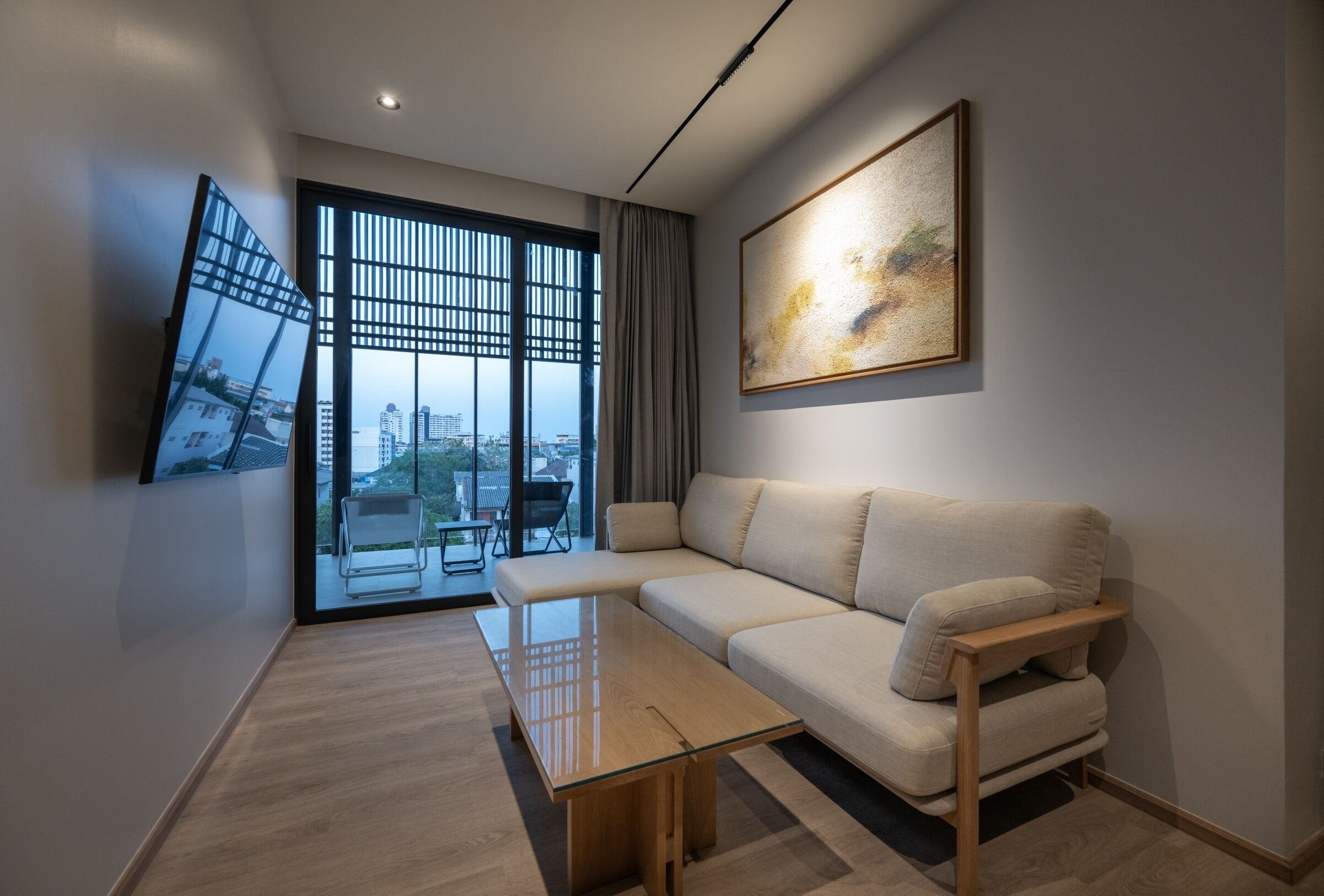 A modern living room with a sofa, a glass coffee table, a TV mounted on the wall, and a large window with a view of buildings.